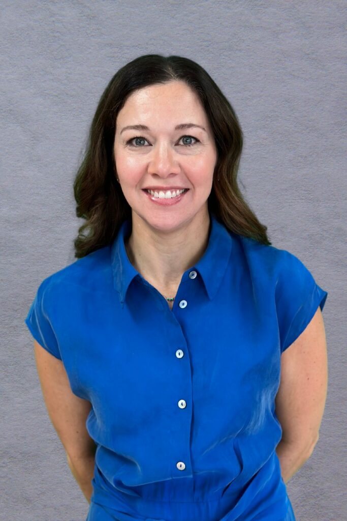 Woman with long black hair in blue shirt standing in front of a gray background.