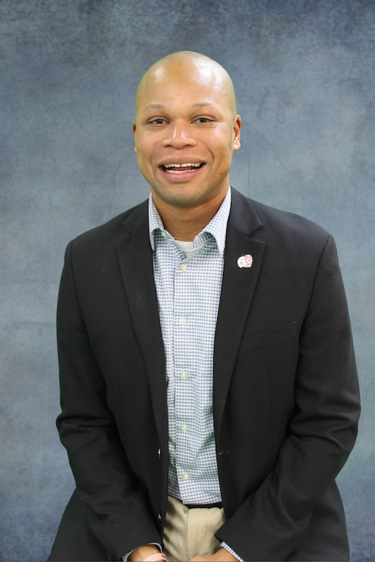 Man in a dark suit jacket with a lapel pin in front of a gray background.