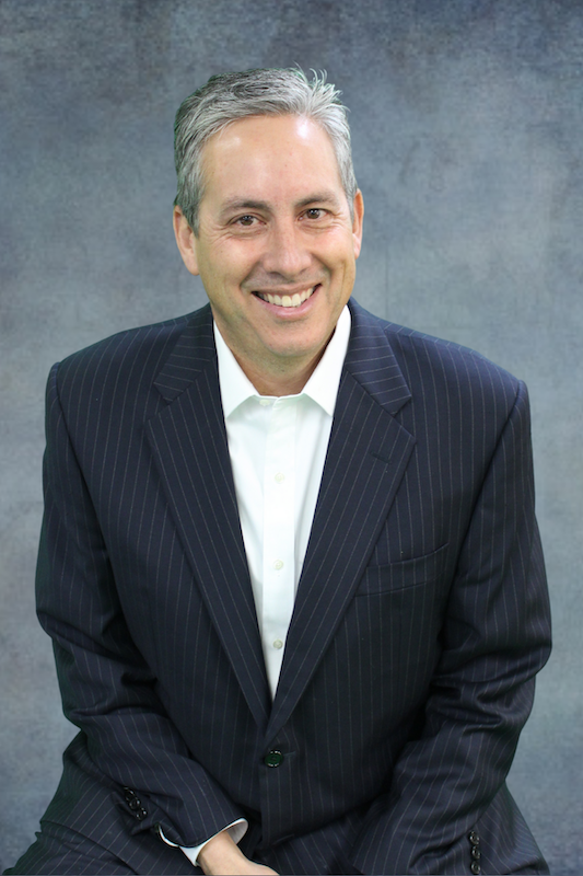 Man in dark suit jacket in front of gray background.
