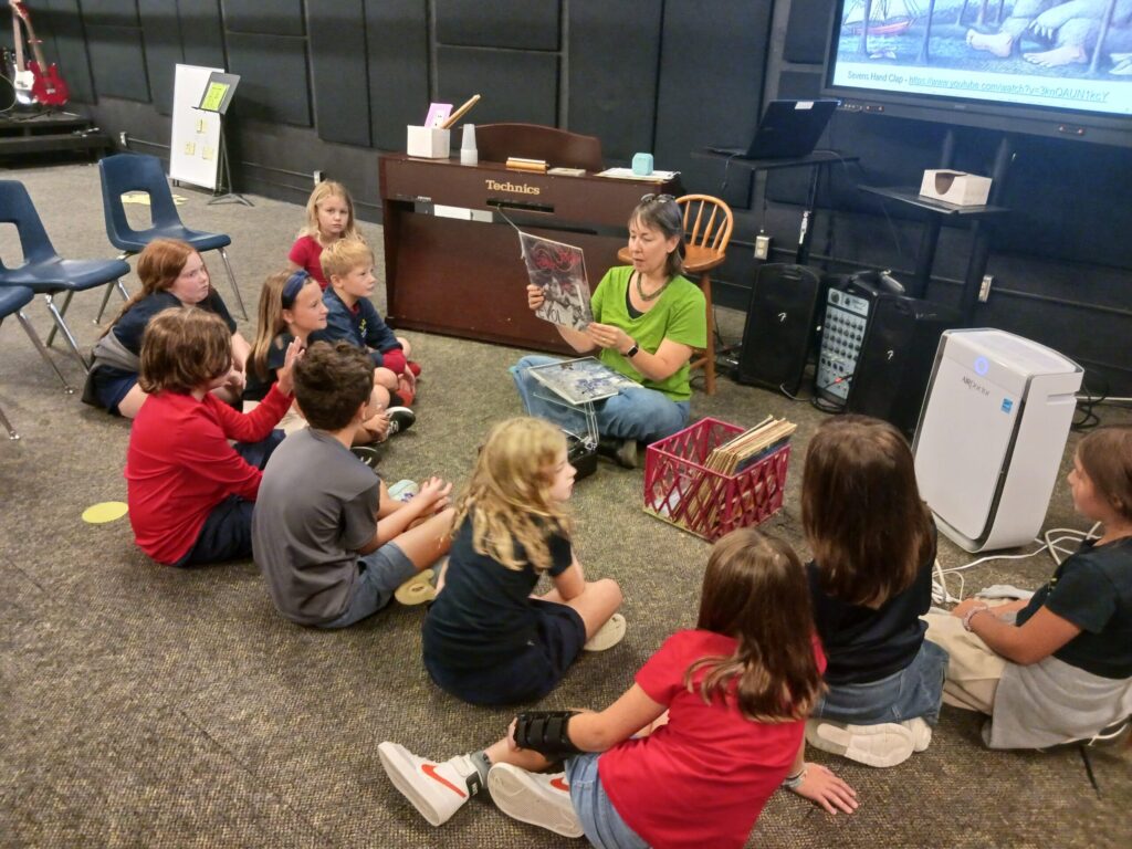 A teacher sitting cross-legged on the floor in a bright classroom, reading a large vinyl record sleeve to a group of young students sitting around her in a circle. In the background are musical instruments including a Technics electronic piano, speakers, and a large display monitor.