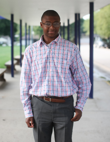 Portrait of a smiling young Black man wearing glasses, a long-sleeved pink and blue plaid button-down shirt, dark gray pinstriped trousers, and a brown belt, standing under a covered outdoor walkway with dark blue support pillars.