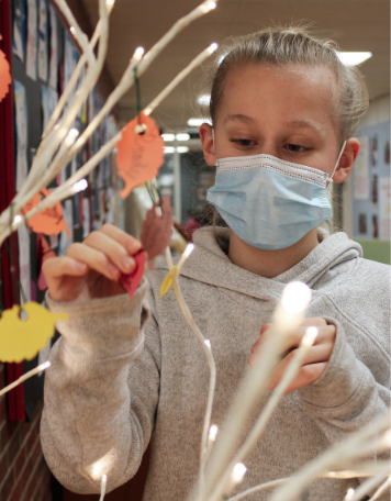 A young girl wearing a light blue surgical face mask and a gray hoodie hanging colorful paper leaf cutouts onto the branches of an illuminated decorative tree display in a school hallway.