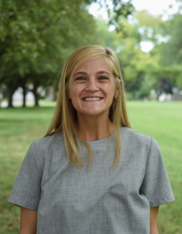 Portrait of a smiling woman with straight, long blonde hair, wearing a short-sleeved gray top, posing outdoors with a bright green lawn and trees in the background.