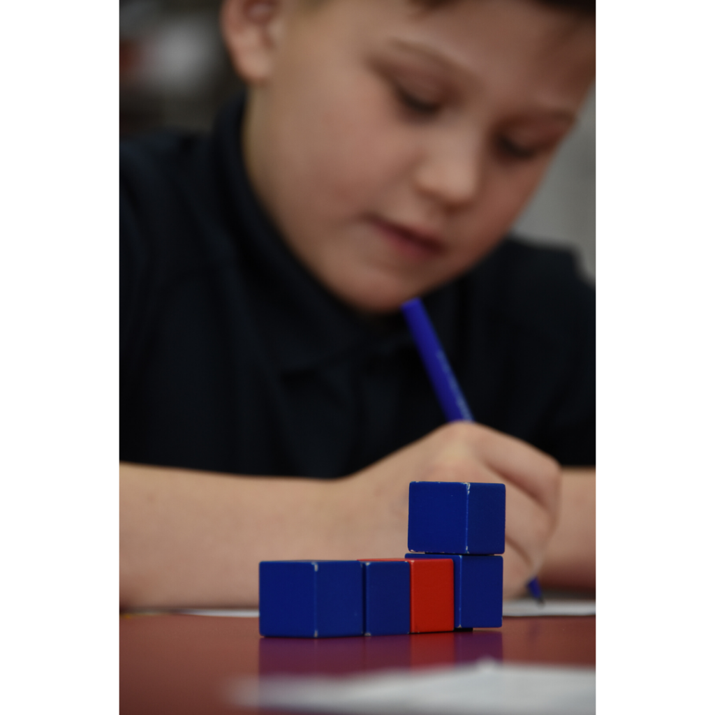 Close-up of small blue and red wooden counting blocks arranged on a table in the foreground, with a young boy writing with a pencil blurred softly in the background.
