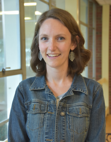Portrait of a smiling woman with shoulder-length wavy brown hair, wearing a medium-wash denim jacket and silver leaf-shaped filigree earrings. She is standing indoors in front of large glass windows and a brick wall background.