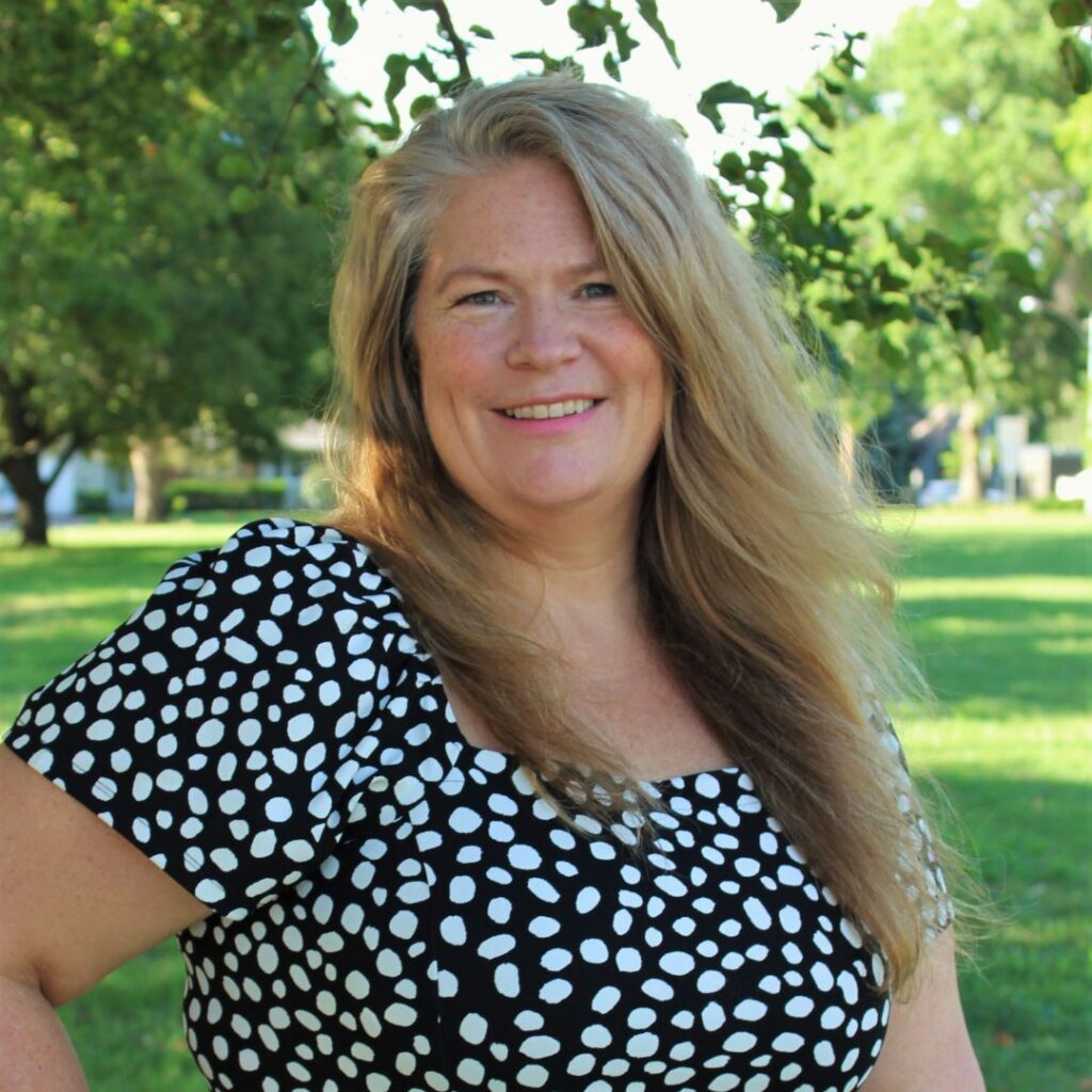 A portrait of a woman with long blonde hair and a warm smile, wearing a black top with white polka dots, posing outdoors in front of a green lawn and trees.
