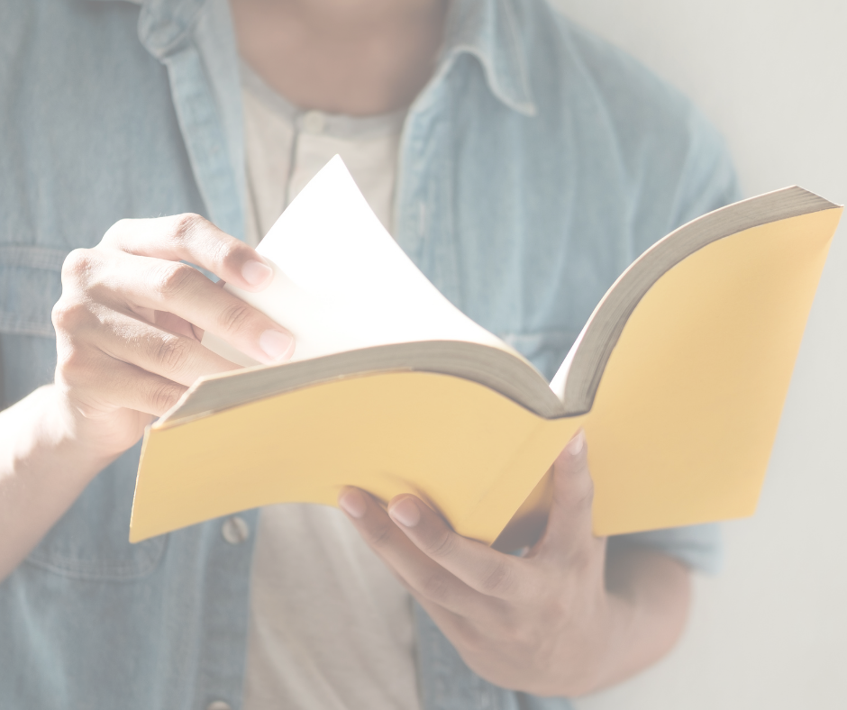 Close-up of a person wearing a light blue denim shirt holding an open book with a yellow cover, gently flipping through its white pages with soft, bright lighting.