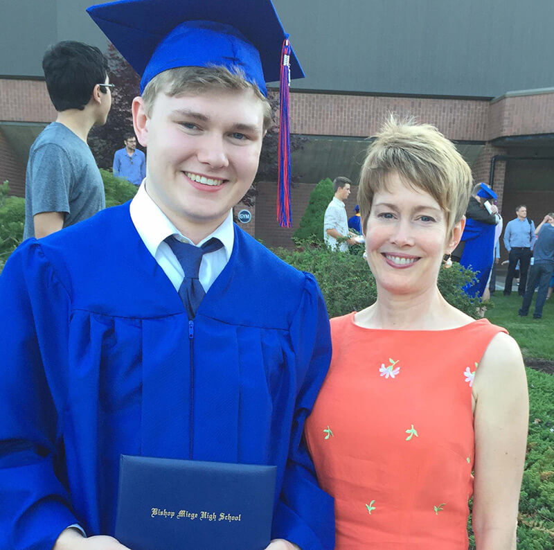 Young graduate in blue cap and gown holding a highschool diploma standing next to an adult woman.