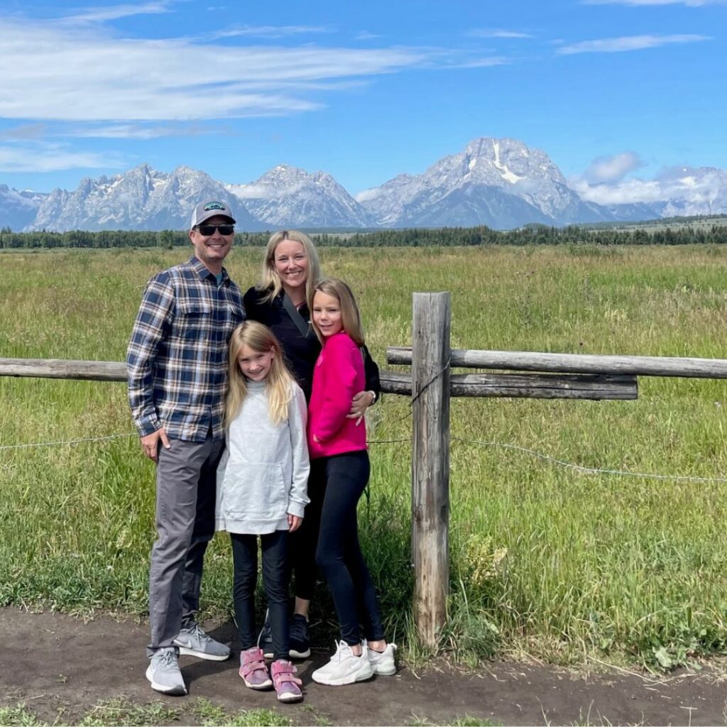 A family of four posing for a picture outdoors in front of a wooden fence. The family consists of two parents standing behind their two young daughters, all smiling. Behind them is an open grassy field with a dramatic, snow-capped mountain range under a partly cloudy blue sky.