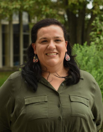 Portrait of a smiling woman with long black hair, wearing an olive green button-up shirt with front flap pockets and dark floral teardrop-shaped earrings, posing outdoors against a lush green garden background.