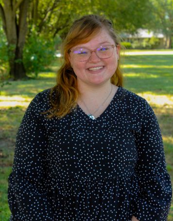 Portrait of a smiling woman with strawberry blonde hair, wearing clear-framed glasses and a long-sleeved black dress with a white polka dot pattern. She is posing outdoors with a sunlit park and trees in the background.
