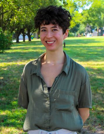 Portrait of a smiling person with short, dark curly hair, wearing an olive green button-up shirt and dangling earrings, posing outdoors in front of a grassy park with sunlit trees.