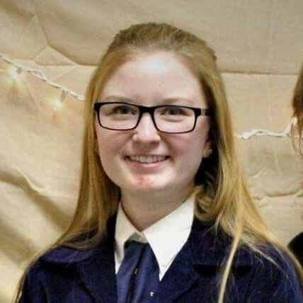 Young student with glasses smiling in front of a wall decorated with string lights.