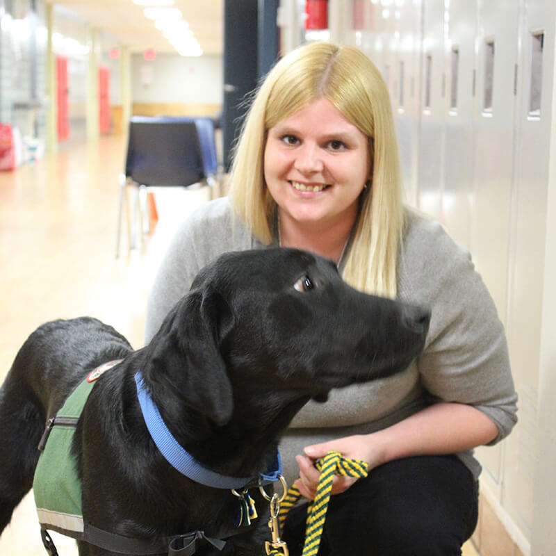 Young person with long blong hair crouched down petting a black dog in front of school lockers.