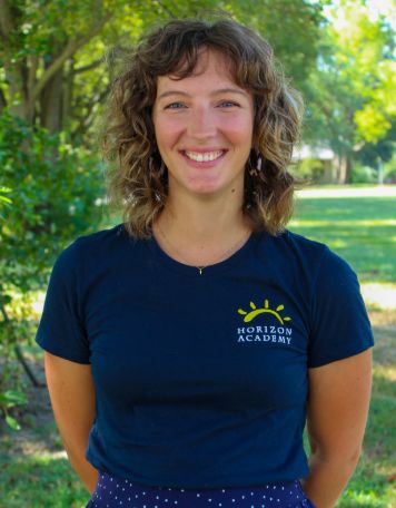 Portrait of a smiling woman with medium-length curly brown hair, wearing a dark blue t-shirt with the "Horizon Academy" yellow sun logo, posing outdoors in front of green trees and grass.