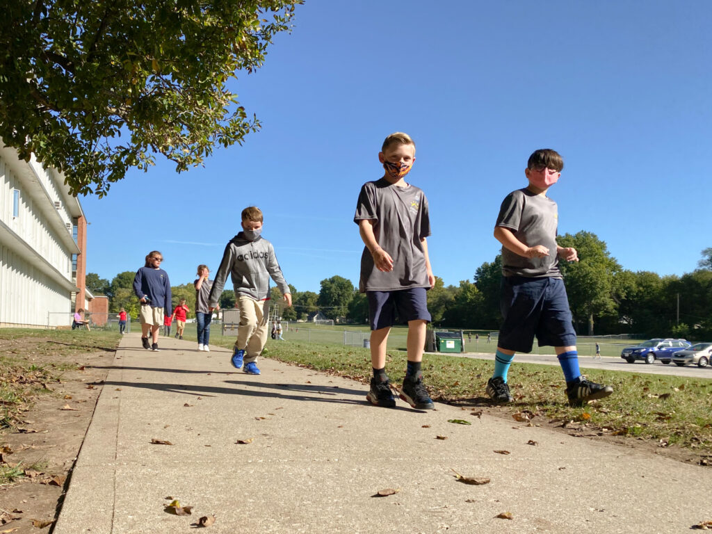 A group of elementary school children wearing face masks walk in a single file line along a concrete sidewalk outside a school building on a bright, sunny day.