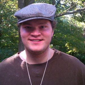 Young man standing outside while wearing a cap and brown shirt.