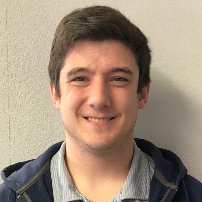 Young man with brown hair smiling in front of a beige wall.