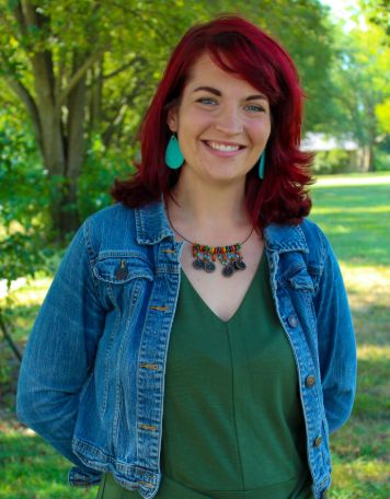 Portrait of a smiling woman with vibrant red shoulder-length hair, wearing a denim jacket over a green v-neck top, turquoise teardrop earrings, and a beaded necklace. She is standing outdoors with a green grassy park and trees in the background.
