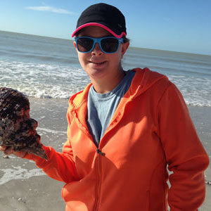 Person on a beach wearing sunglasses and an orange jacket while holding a conch shell.