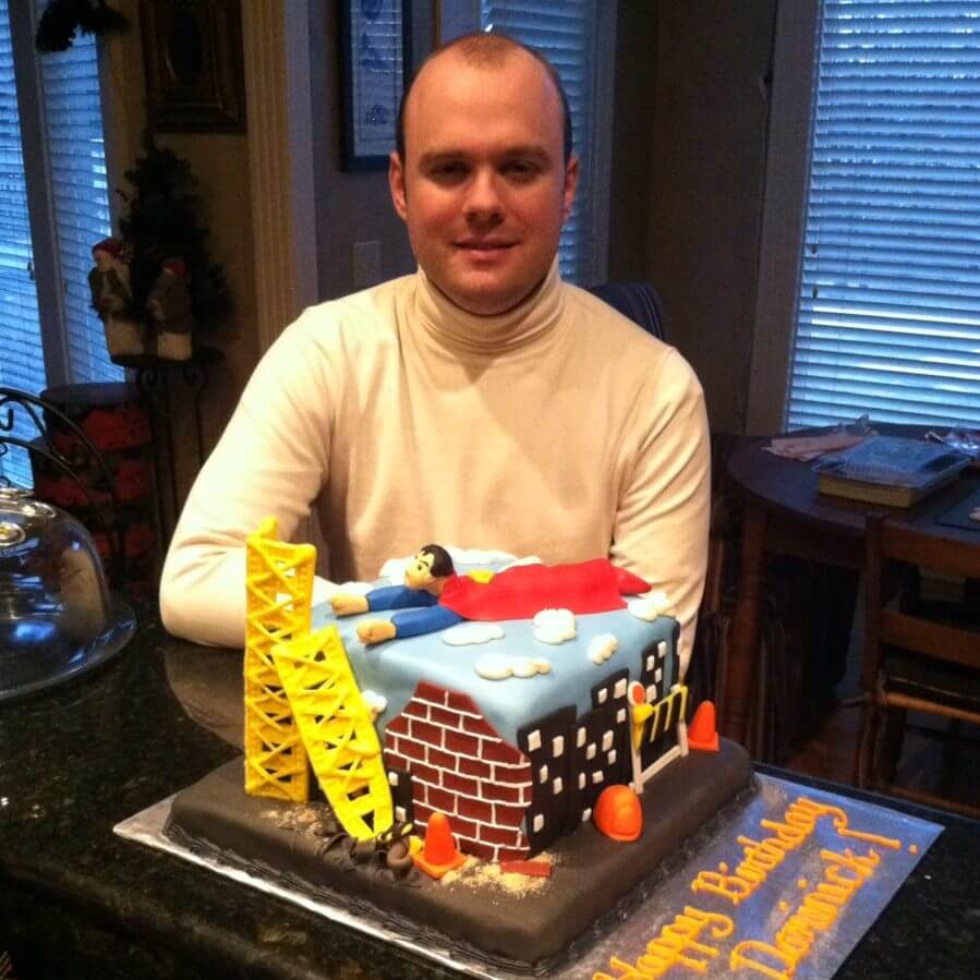 Person in a white turtle neck sitting by a Superman birthday cake.