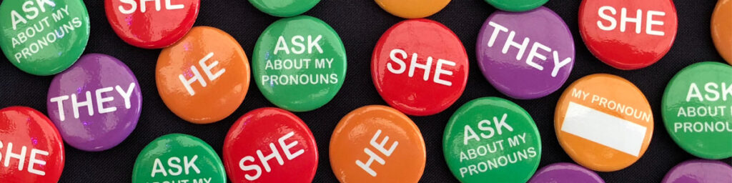 Wide banner image filled with various colorful round pin-back buttons on a dark fabric background. The buttons come in red, green, orange, and purple, displaying text such as "SHE", "HE", "THEY", "ASK ABOUT MY PRONOUNS", and a blank orange button with space to write custom pronouns.