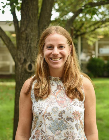 Portrait of a smiling woman with shoulder-length wavy blonde hair, wearing a sleeveless floral high-neck blouse in muted pastel tones, standing outdoors in front of a tree and green lawn.