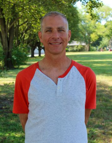Portrait of a smiling man with short graying hair, wearing a light gray henley shirt with orange raglan sleeves, posing outdoors in front of a green grassy park and trees.