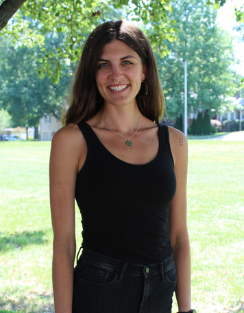 Portrait of a smiling woman with long brown hair, wearing a black tank top, dark jeans, a nose ring, and a small necklace, posing outdoors with a sunny green park in the background.