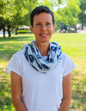 Headshot of a smiling woman with short dark hair and a patterned scarf standing outdoors in a park setting.