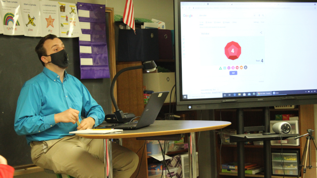 A male teacher wearing a face mask, light blue button-down shirt, and khakis sits at a desk in a classroom holding a pencil. Beside him is a laptop connected to a large interactive display screen showing a red digital die rolling a 4.