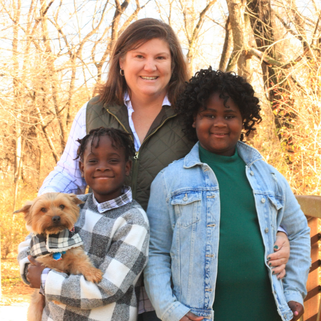 An outdoor family portrait featuring a smiling woman, two young Black children, and a small tan dog. The woman stands in the center wearing an olive green vest over a white collared shirt. On the left, a child in a black-and-white checkered sweater holds the small dog, who wears a matching plaid collar accessory. On the right, a child with curly hair wears a green top and a light blue denim jacket. Bare autumn trees fill the background.