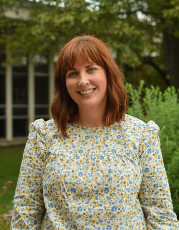Portrait of a smiling woman with shoulder-length reddish-brown hair and front bangs, wearing a long-sleeved floral blouse with blue and yellow flowers, standing outdoors with lush green foliage in the background.
