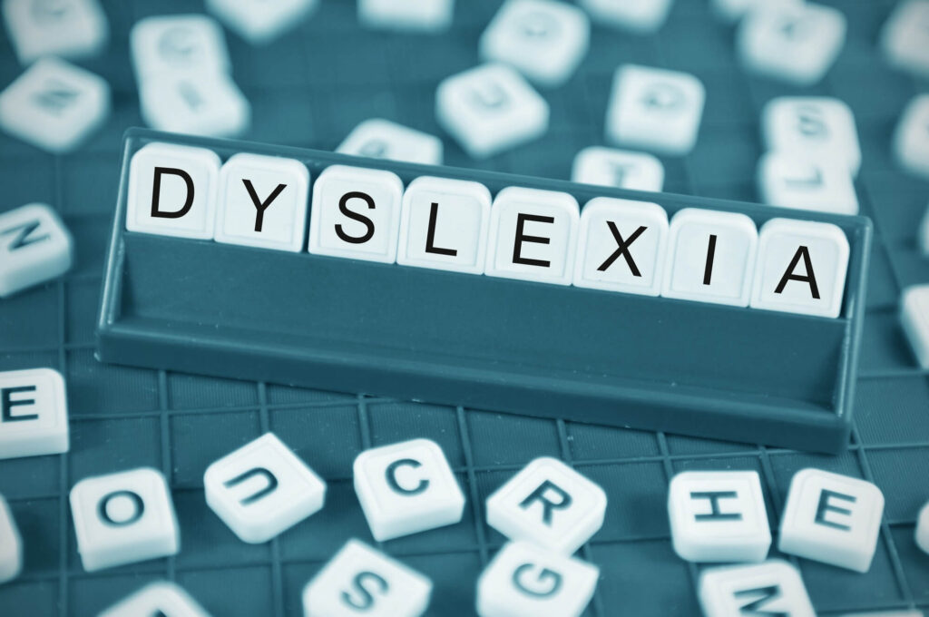 Close-up photograph with a teal filter showing letter tiles arranged in a tile rack to spell out the word "DYSLEXIA." Other scrambled letter tiles are scattered around the game board in the blurred background and foreground.