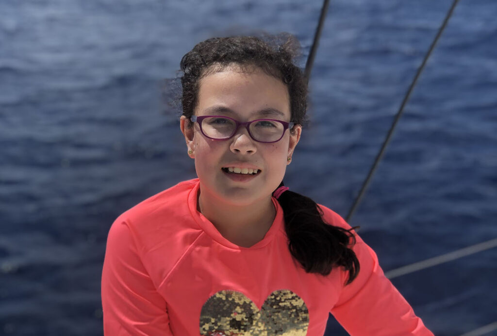 Portrait of a young girl wearing purple-rimmed glasses and a bright coral long-sleeved shirt with a gold sequin heart on the front. Her dark hair is pulled back into a side ponytail, and she smiles against a background of deep blue open water, likely on a boat.