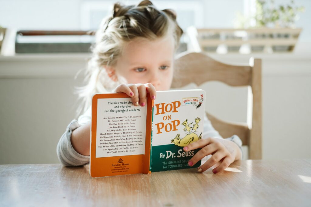 A young girl sitting at a wooden table reading the Dr. Seuss board book "Hop on Pop". She holds the book open in front of her face, displaying both the back cover list of titles and the front cover.