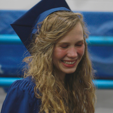 Smiling graduate with long hair wearing a blue cap and gown.
