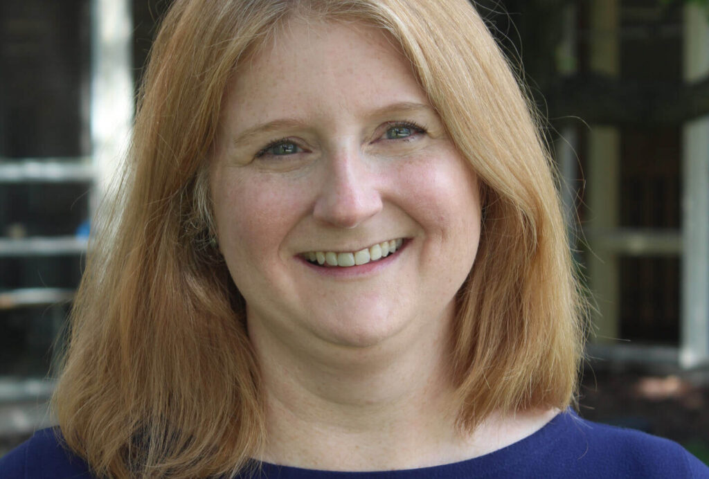 Close-up headshot of a smiling woman with straight, shoulder-length blonde hair and fair skin with freckles, wearing a dark blue top.