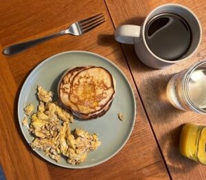 Overhead view of pancakes and scrambled eggs on a gray plate, with coffee, water, and orange juice on a wooden table; a casual breakfast.