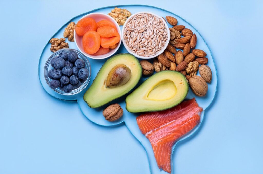 A brain-shaped arrangement depicts blueberries, apricots, almonds, grains, avocado, walnuts, and salmon on blue, suggesting foods for a brain-healthy diet.
