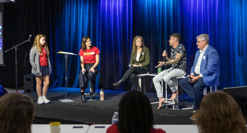 A four-person panel discusses question asked by Horizon Academy student onstage before blue curtains; a man holds a microphone to give his answer.