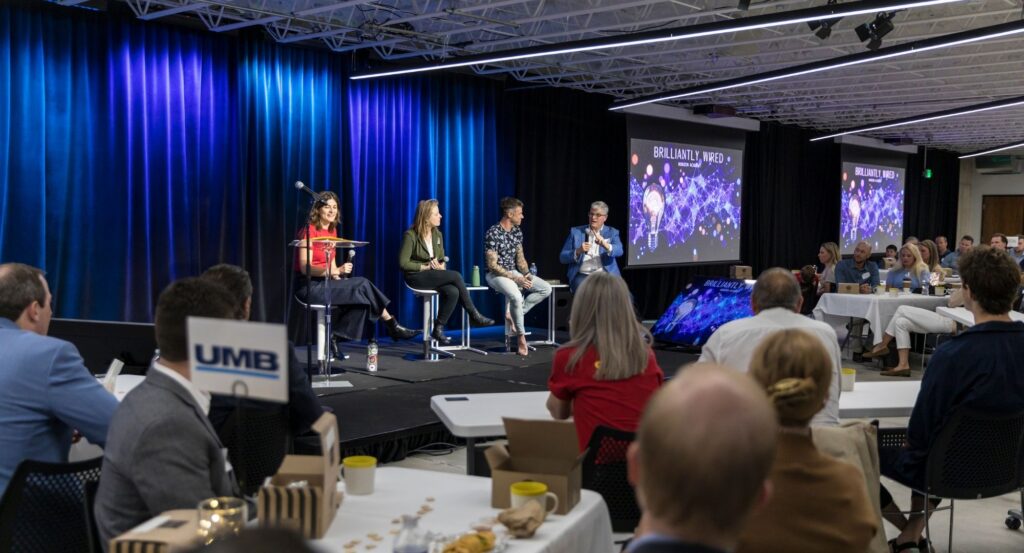 Three Brilliantly Wired speakers and panelist sit on a raised conference stage against blue curtains as attendees watch from tables; screens show a colorful event graphic, creating an engaged atmosphere.