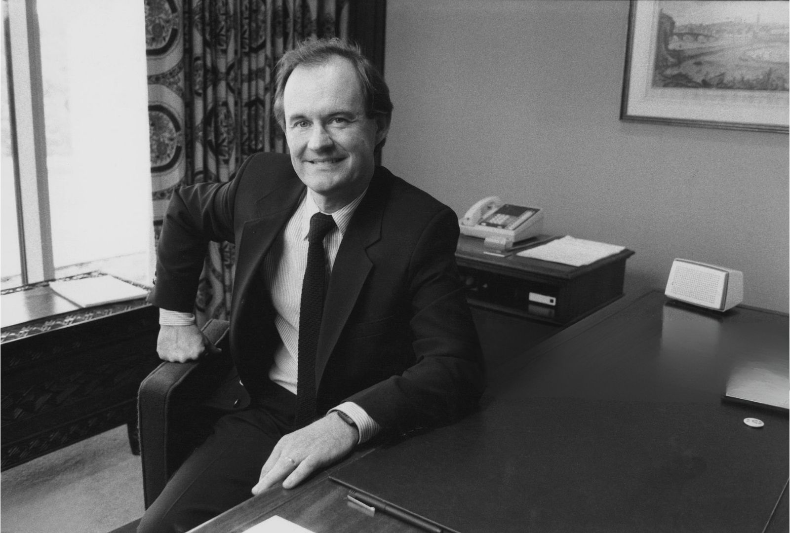 A black-and-white portrait of David Boies in a suit seated beside a large office desk, with papers, a telephone, and framed art behind him.