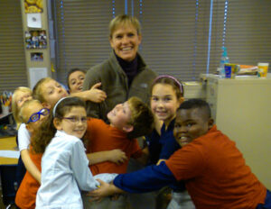 Smiling teacher surrounded by schoolchildren in a playful group hug in a classroom, conveying warmth, affection, and cheerful camaraderie.