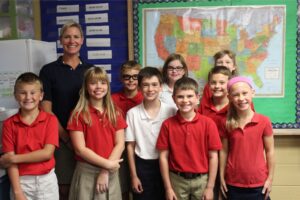 A smiling teacher poses with eight schoolchildren in a classroom, with a colorful U.S. map behind them; the group’s bright expressions convey warmth and pride.